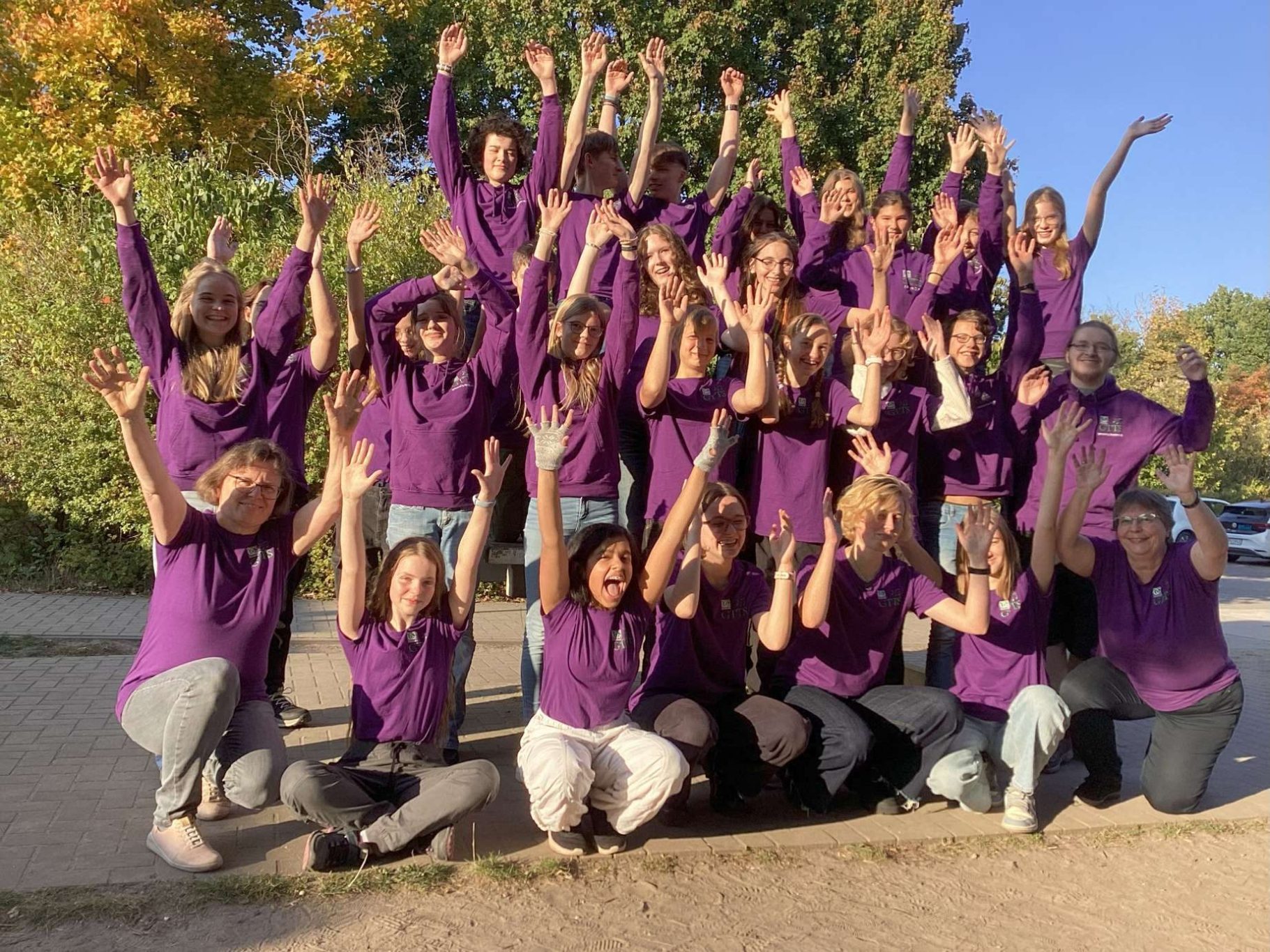 gtts_chor_humboldtgymnasium_gifhorn_foerderverein__neue t-thirts Gruppenfoto von etwa 20 Personen unterschiedlichen Alters, die gemeinsam im Freien stehen und sitzen. Alle tragen violette GTTS T-Shirts